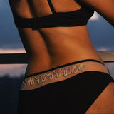 Woman in a black bikini standing against a scenic background with mountains and sky.