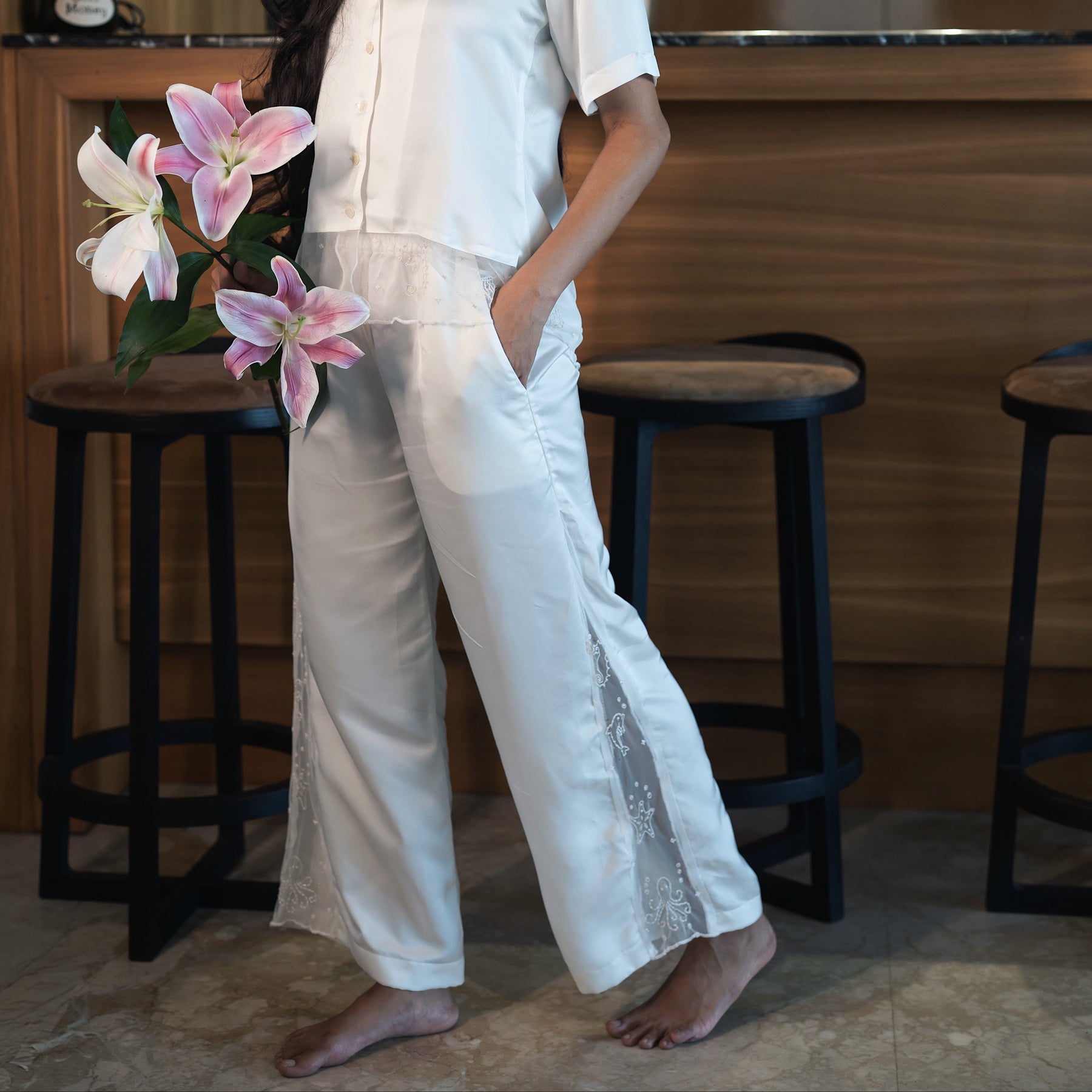 Woman in a white outfit holding pink flowers in a room with wooden furniture.