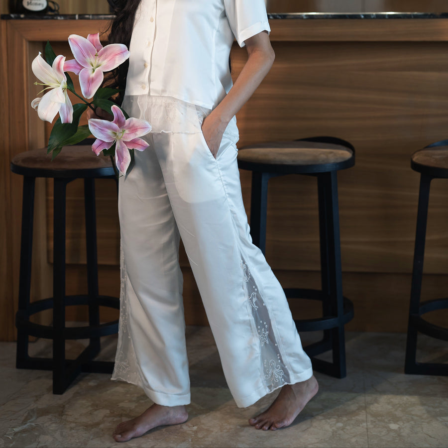 Woman in a white outfit holding pink flowers in a room with wooden furniture.