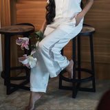 Woman in a white outfit sitting on a stool with flowers, in a room setting.